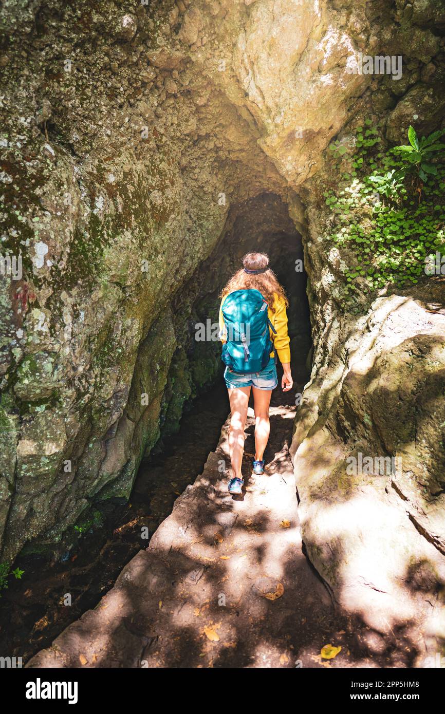 Description: Tourist woman entering a tunnel along an overgrown jungle trail next to a canal in Madeira rainforest. Levada of Caldeirão Verde, Madeira Stock Photo