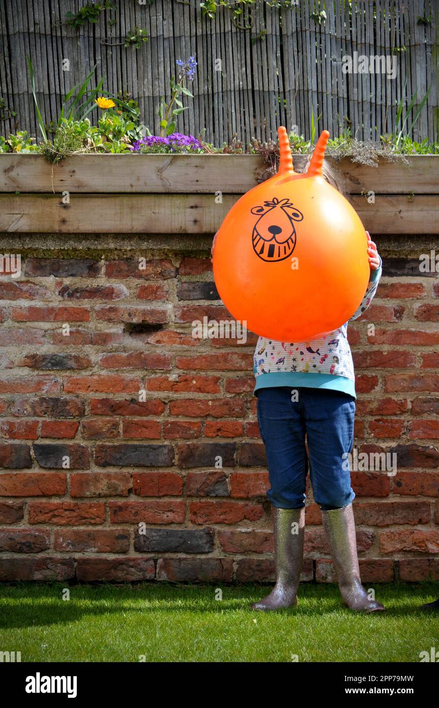 A young girl holding an orange space hopper in front of her face set against a brick garden wall Stock Photo
