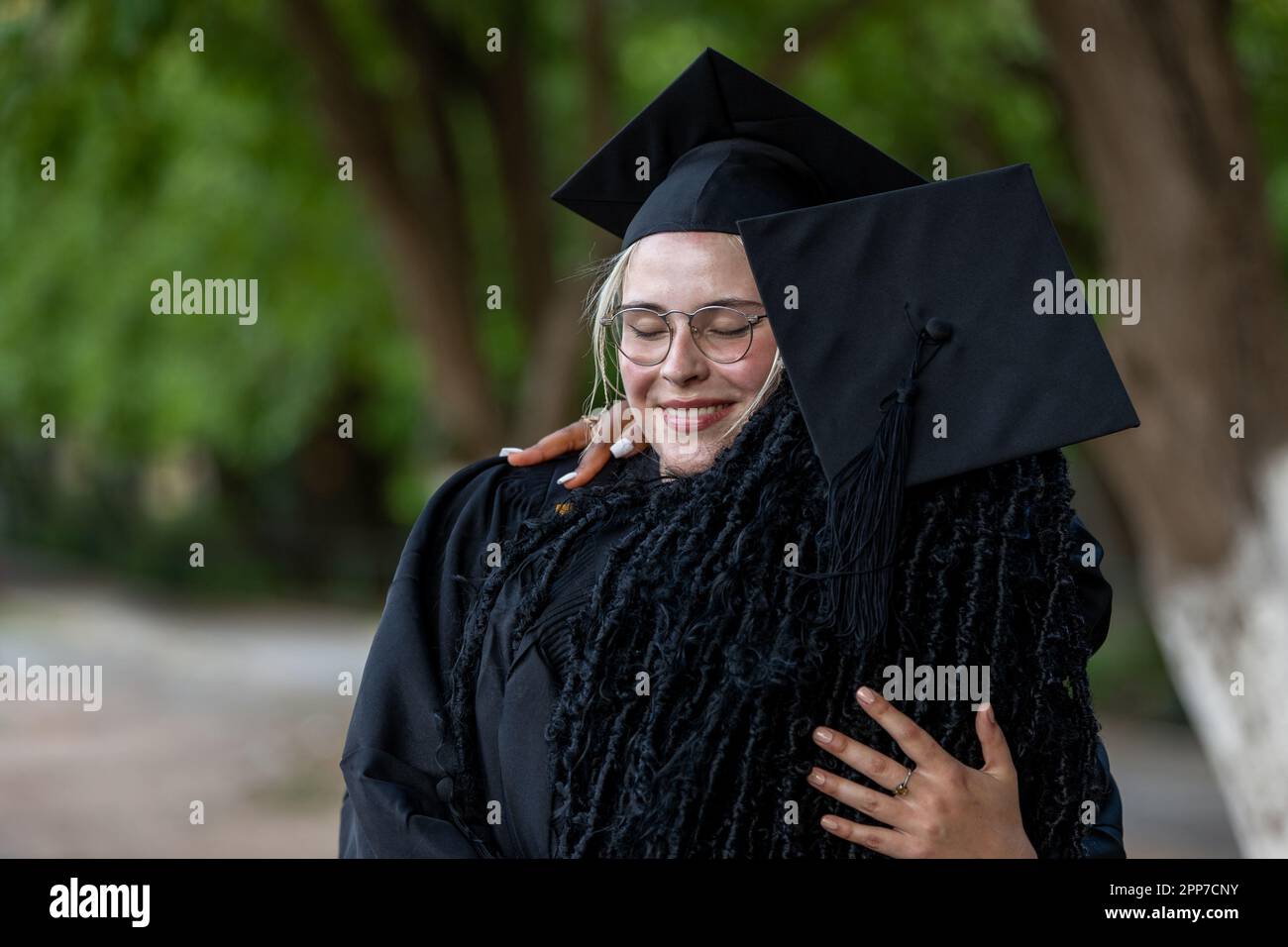 Caucasian Female and Black European College Graduates wearing caps and gowns hug after graduation, proud of academic success, degrees and gaining know Stock Photo