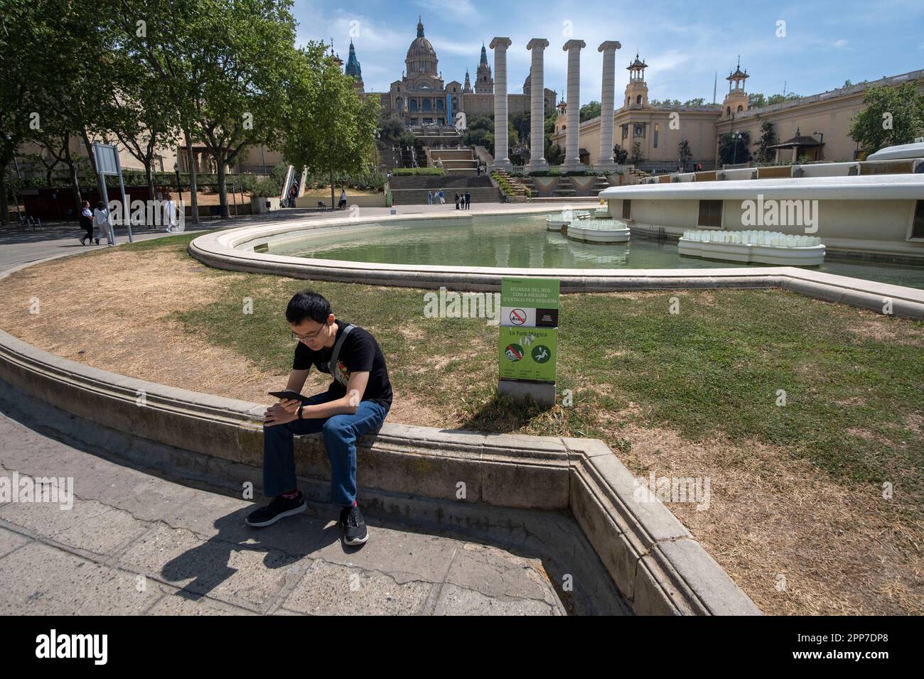 A tourist is seen sitting next to the informative poster of the pump stop as a measure to save water due to drought in La Fuente Mágica. After 30 months without rain in Catalonia and with water reserves at historic lows, the Government of Catalonia has been forced to take water-saving measures by closing the fountains at La Fuente Mágica in Plaça de Josep Puig i Cadafalch which constitutes in itself a great tourist spectacle of water, light and sound. Stock Photo