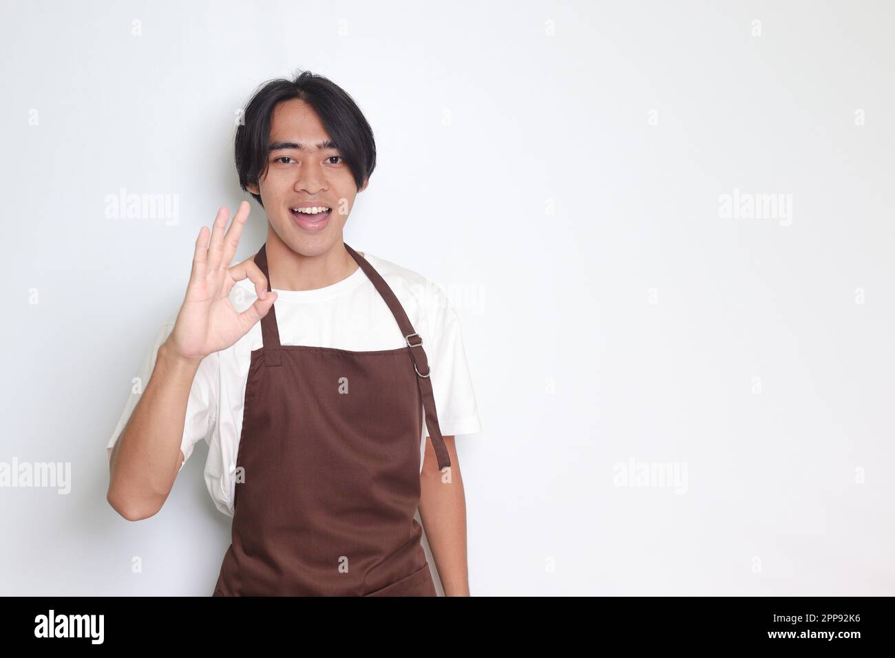 Portrait of attractive Asian barista man in brown apron showing ok hand gesture and smiling looking at camera. Advertising concept. Isolated image on Stock Photo