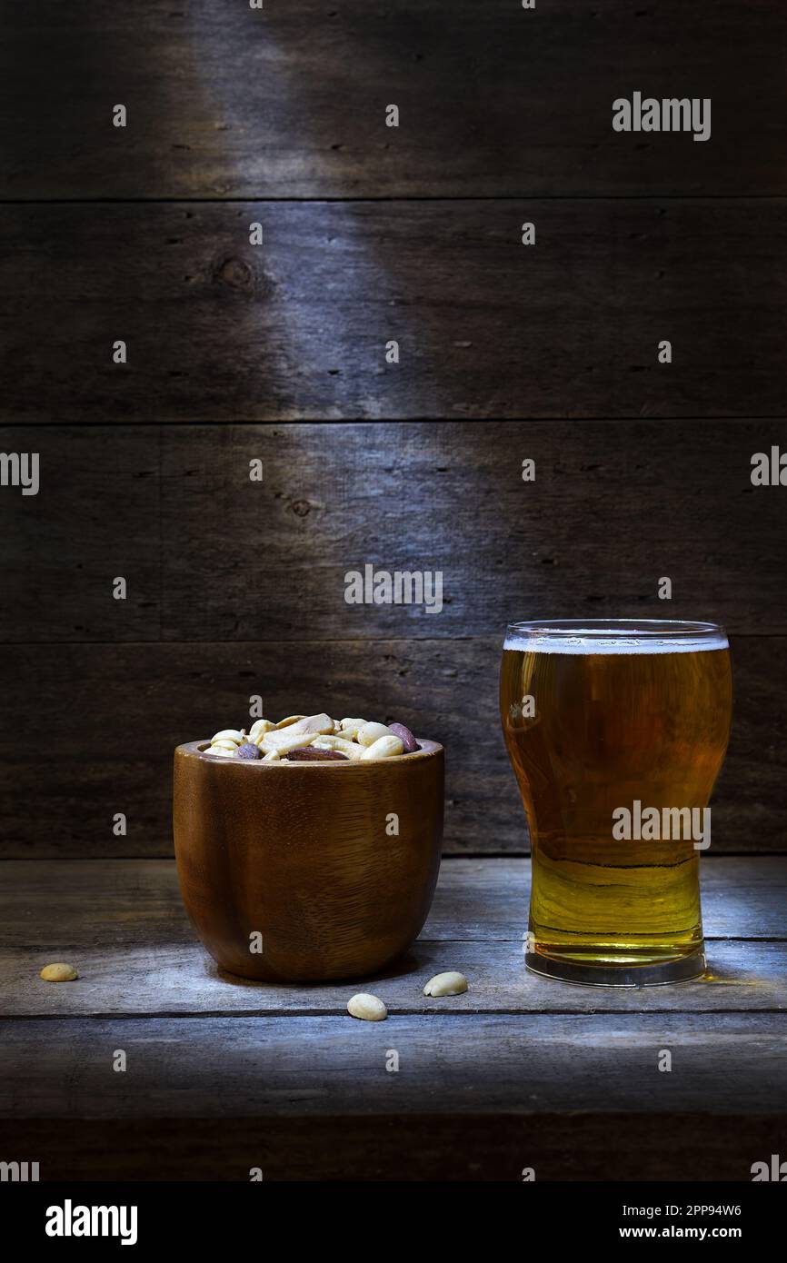 A brown, wooden bowl full of unsalted mixed nuts and a glass of beer in a rustic wooden setting, backlit in a pool of mood lighting; copy space at top Stock Photo