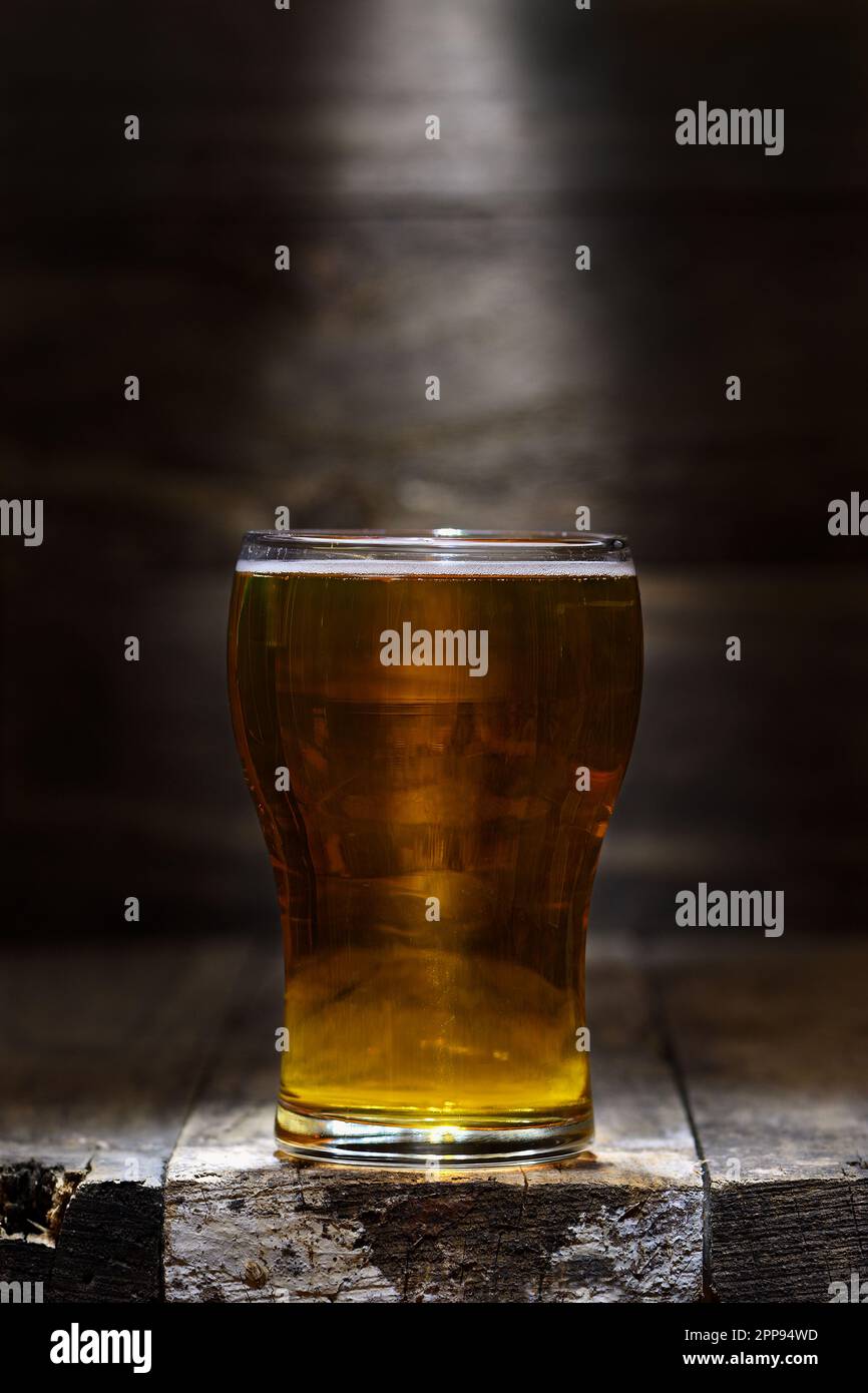 A glass of pale lager beer in a rustic wooden setting, backlit in a pool of mood lighting with copy space to the top of frame; captured in a studio Stock Photo