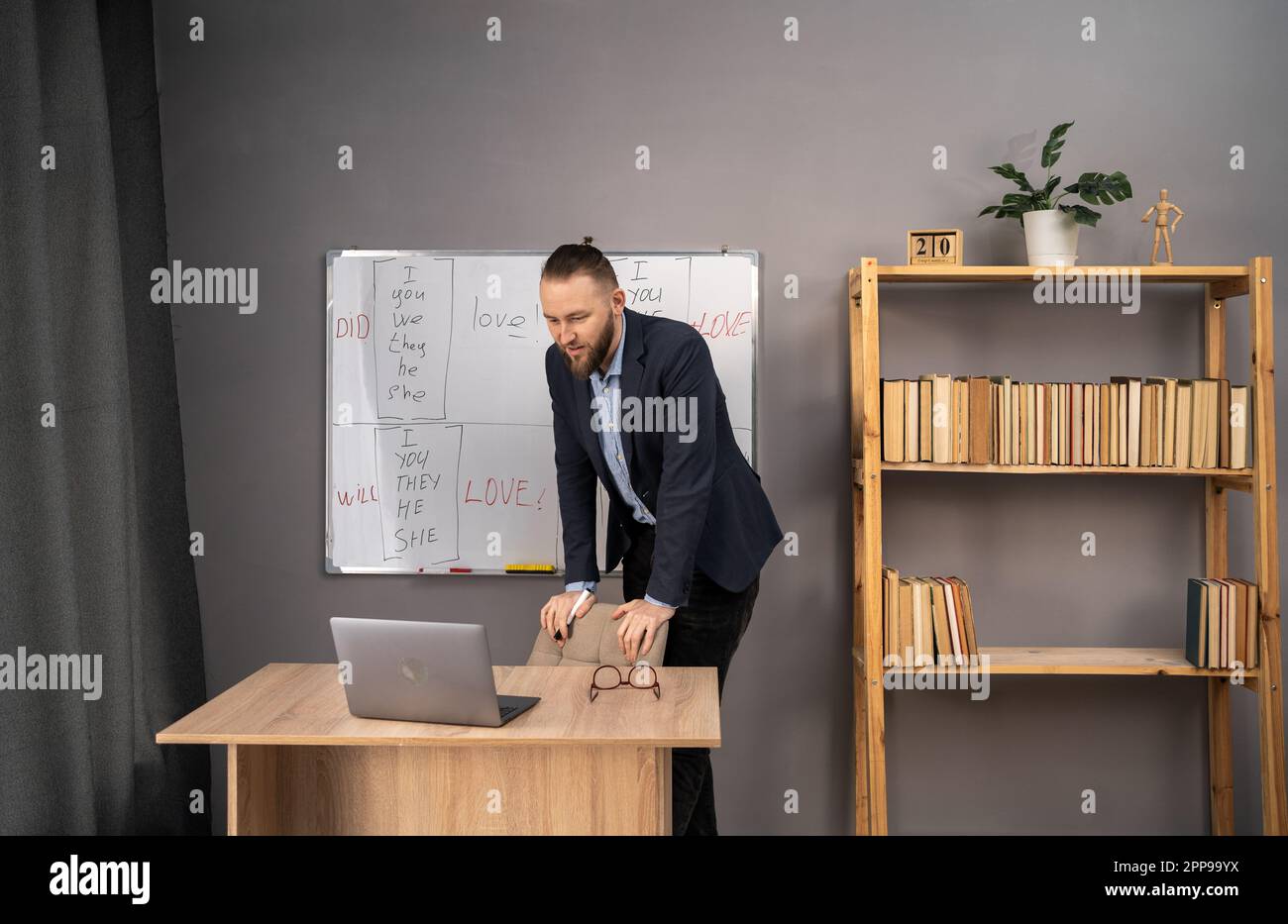 Remote education concept. Happy male teacher giving online English lesson using laptop and blackboard with grammar rules at home office. Stock Photo