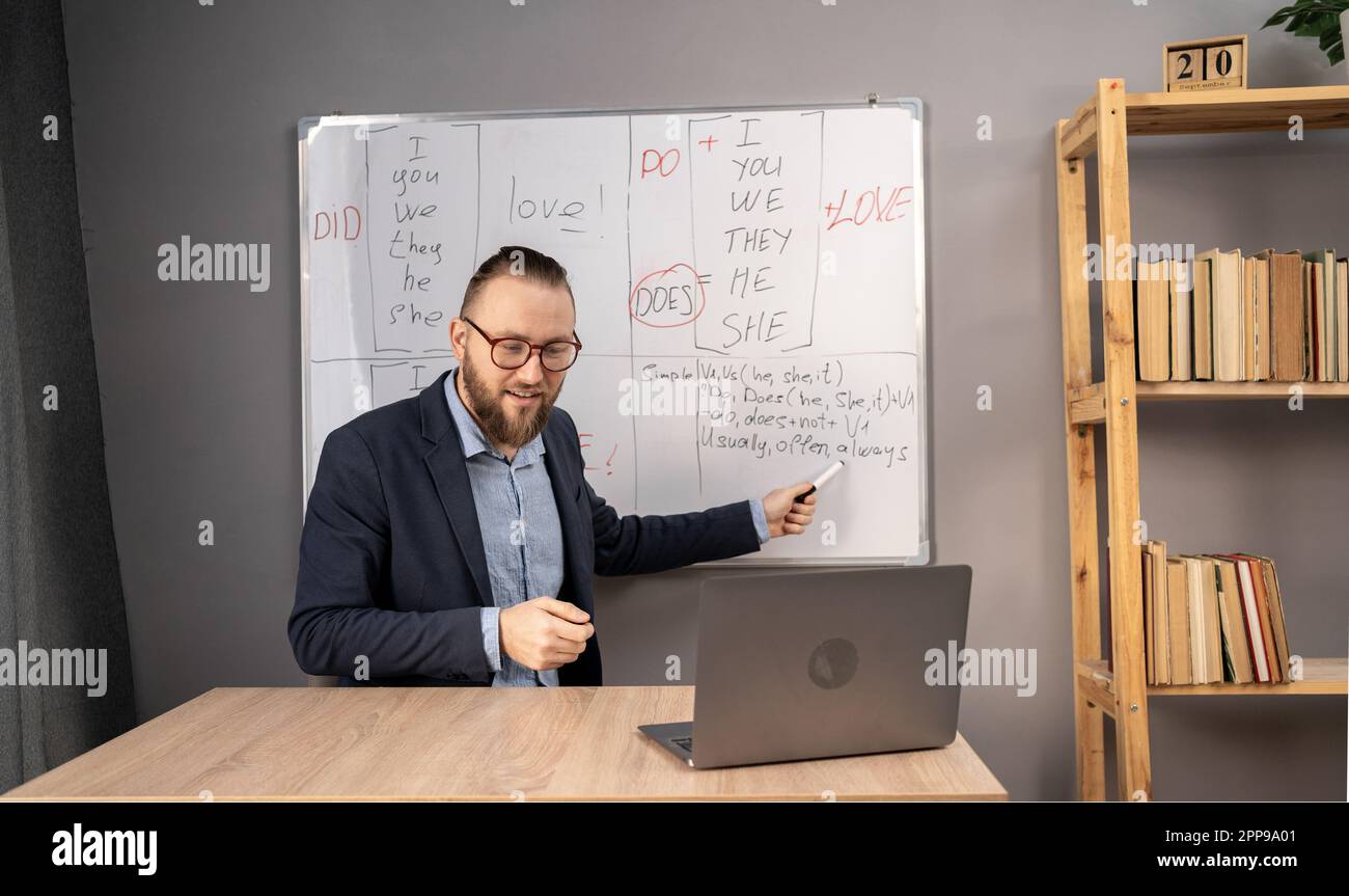 Portrait of smiling adult male lecturer teaching English pointing with marker at grammar rules on whiteboard, watch on students in his online class Stock Photo