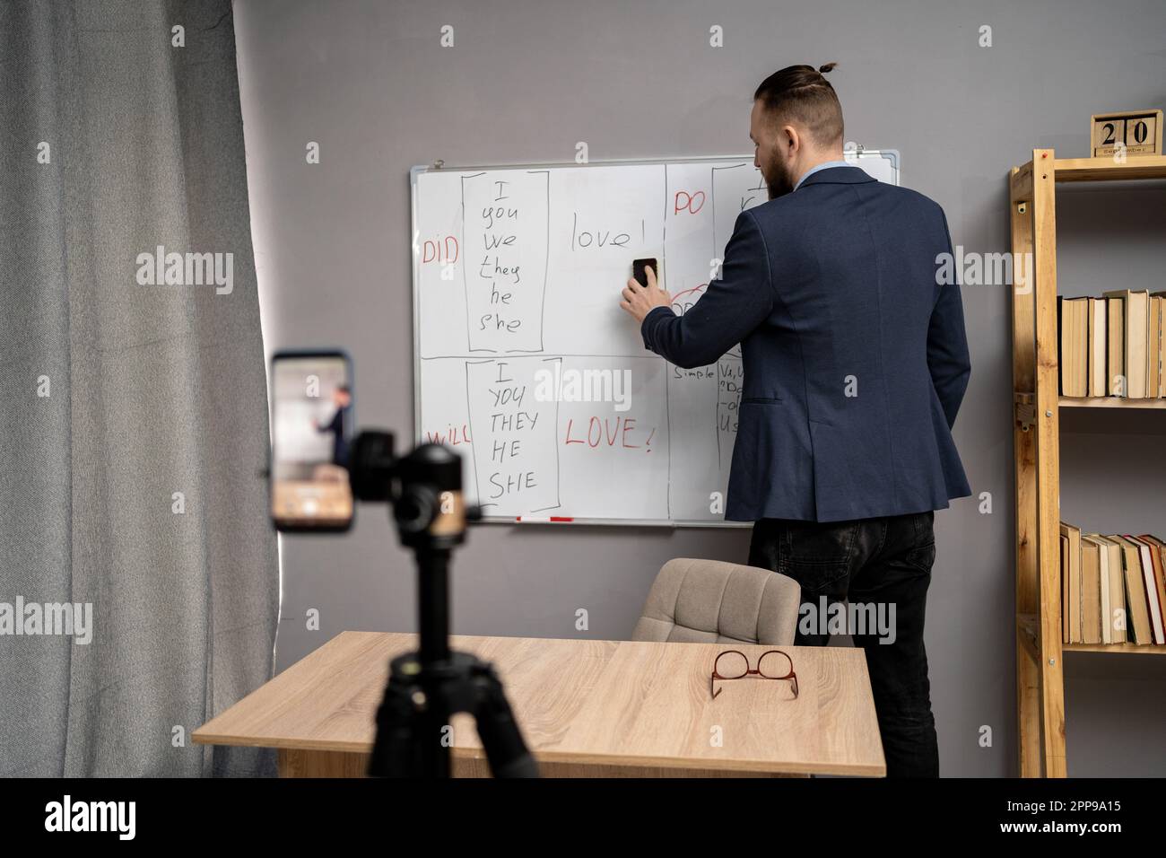 Male teacher standing near blackboard with English grammar rules and having online lesson. Millennial tutor giving foreign language class on video Stock Photo