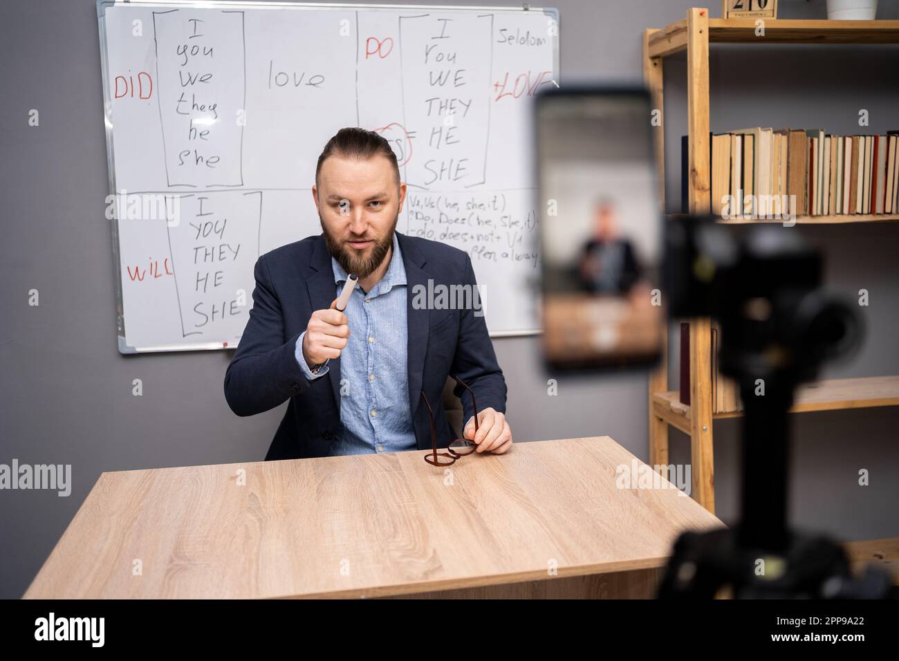 Male teacher giving online English lesson, using smartphone, pointing at webcam sitting before blackboard with grammar rules at home office. Remote Stock Photo