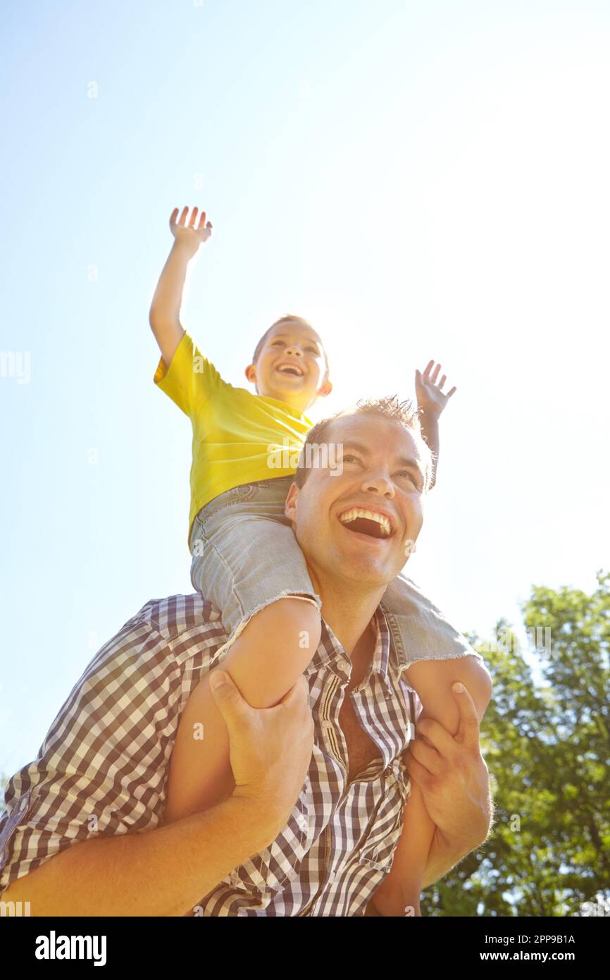 Happy together in the sunshine. Low angle shot of a cute young boy smiling in the sunshine and riding on his dads shoulders. Stock Photo