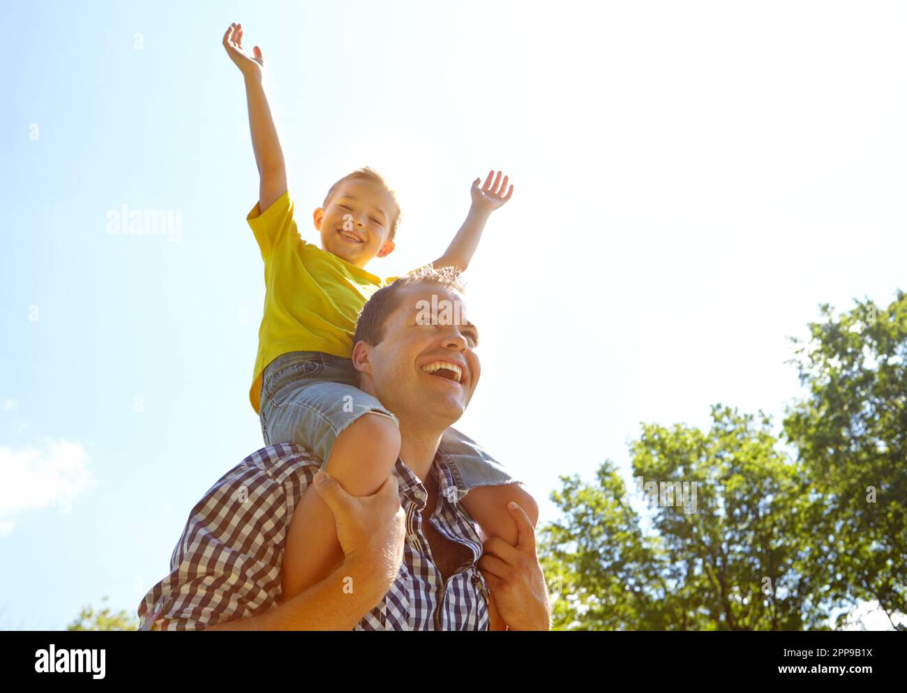 Yay. Low angle shot of a cute young boy smiling widely while riding on his dads shoulders outdoors. Stock Photo