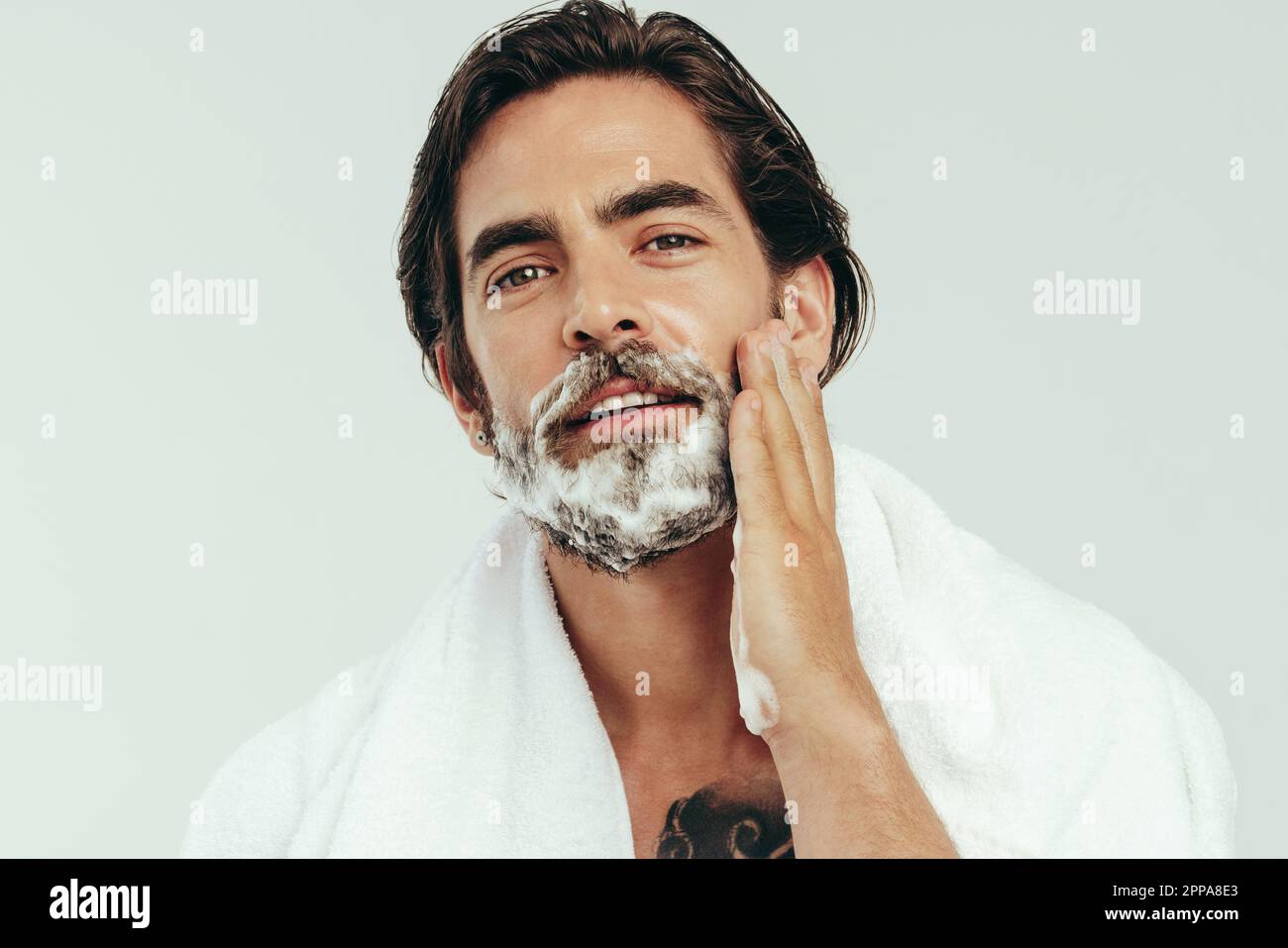 Young man appliying shaving cream to his beard, preparing himself for a smooth and comfortable shave. Man practicing his grooming routine, taking care Stock Photo
