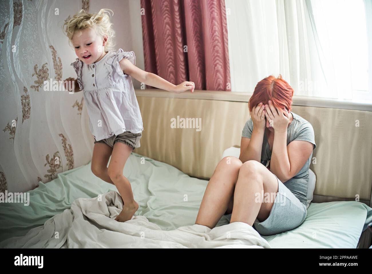 cheerful daughter near sad mother. happy girl jumps on bed next to outbreaks of bad mood mother. woman sits in morning near window with eyes closed wi Stock Photo