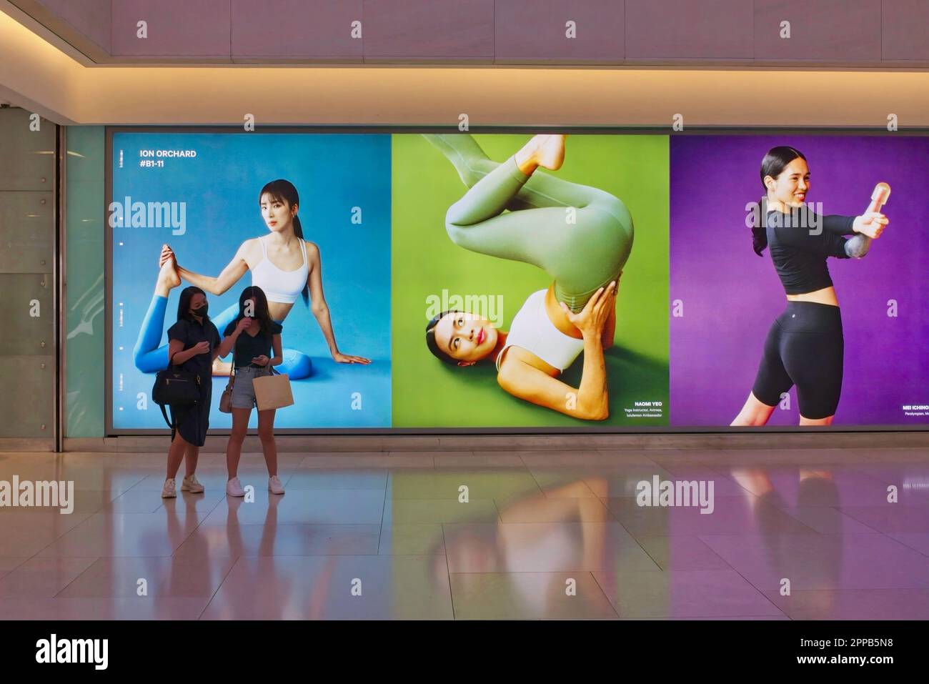 Shoppers at ION Orchard (mall) in Orchard Rd, Singapore pass in front of large yoga-themed advertisements for co. 'lululemon' selling athletic apparel Stock Photo