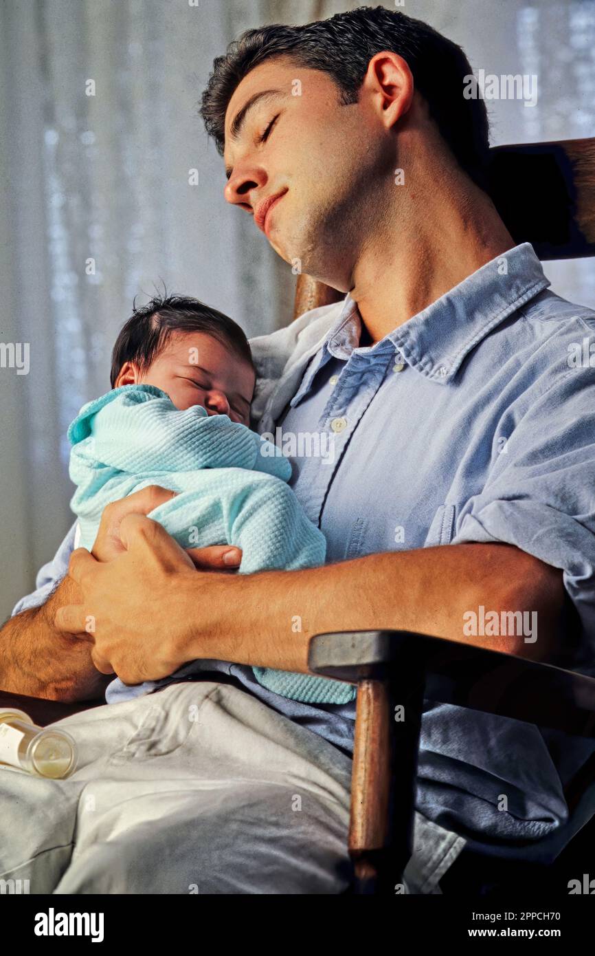 Exhausted, sleep deprived new call Caucasian father holding his baby while sleeping in a rocking chair Stock Photo