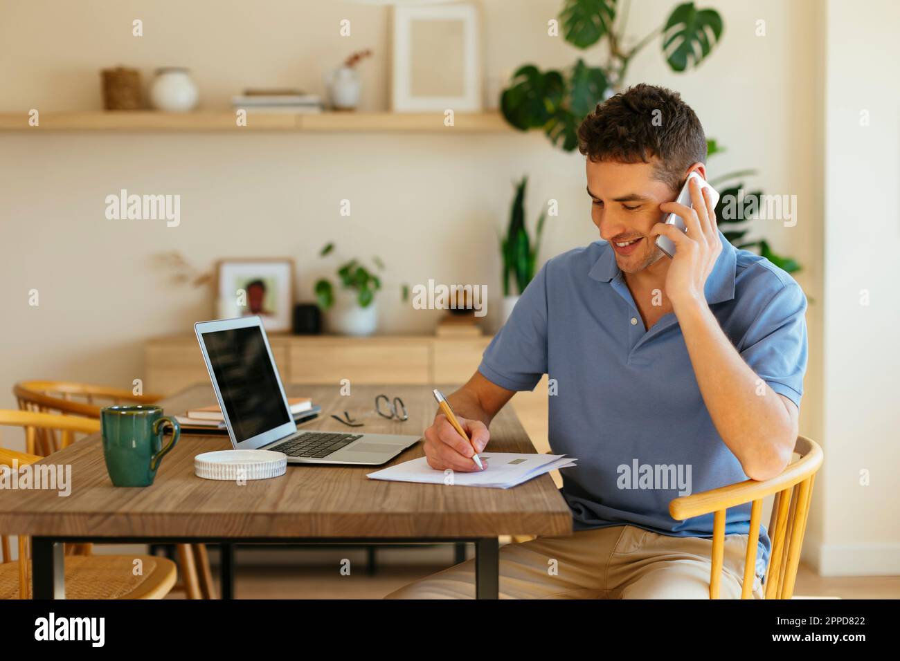 Smiling freelancer talking on mobile phone and taking notes on paper at table Stock Photo