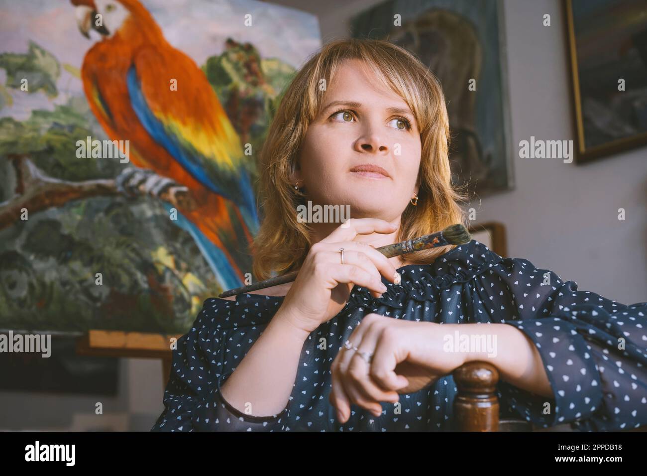 Thoughtful woman with hand on chin holding paintbrush in front of bird painting at workshop Stock Photo