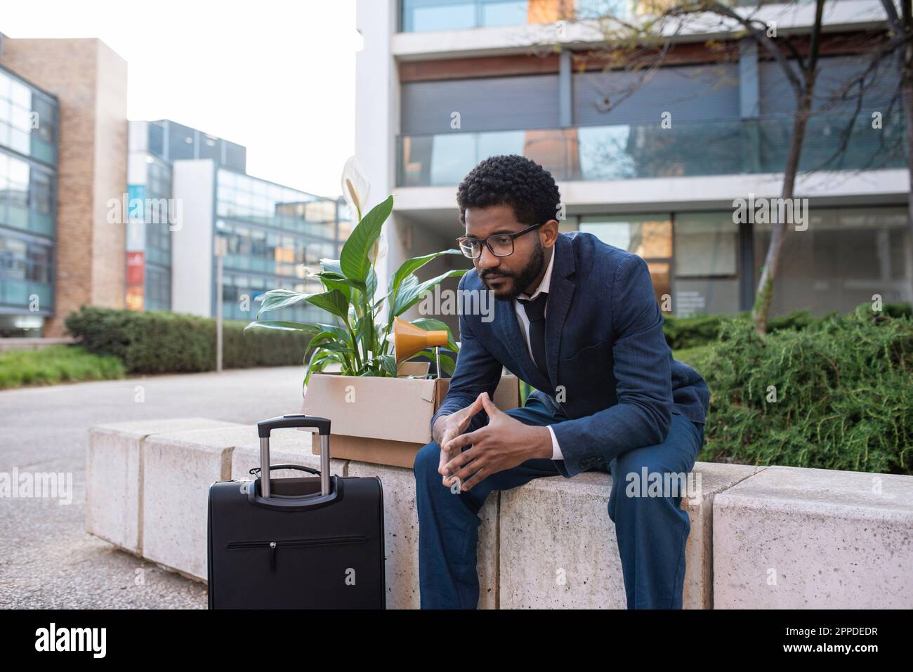 Depressed businessman with suitcase and his belongings sitting outside office building Stock Photo