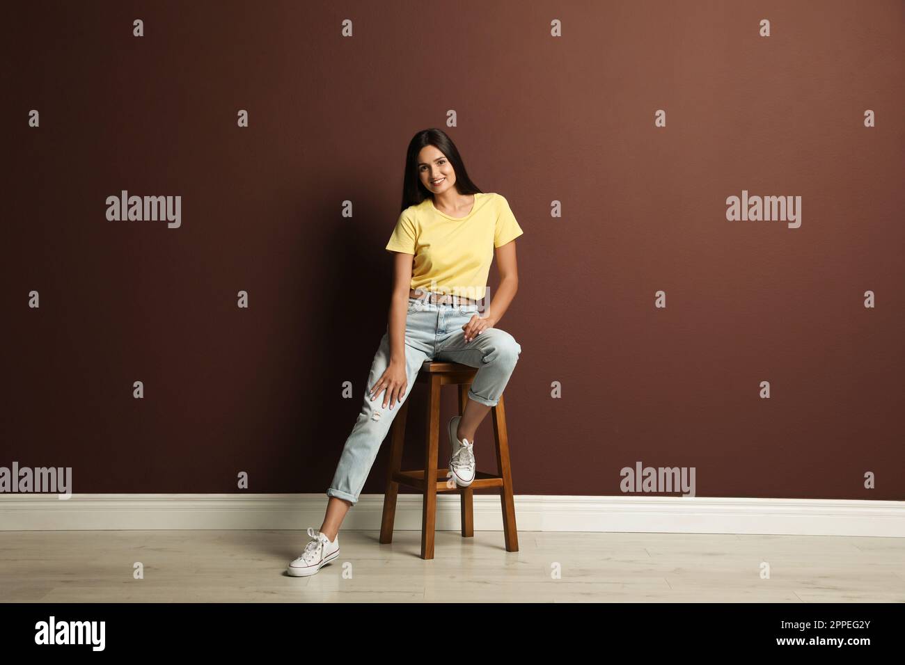 Beautiful young woman sitting on stool near brown wall Stock Photo