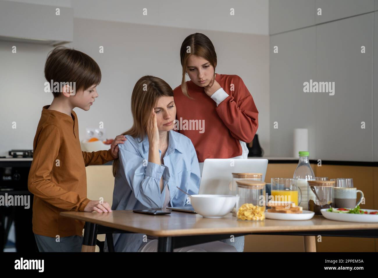 Mom with headache working on laptop on kitchen with children standing
