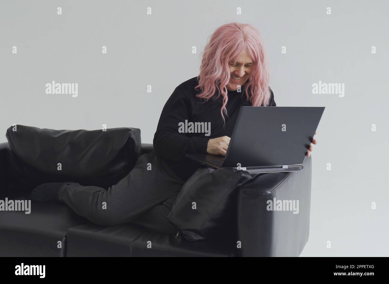 Portrait of a man in a pink wig, sitting on a sofa, typing on a laptop. White background. Stock Photo
