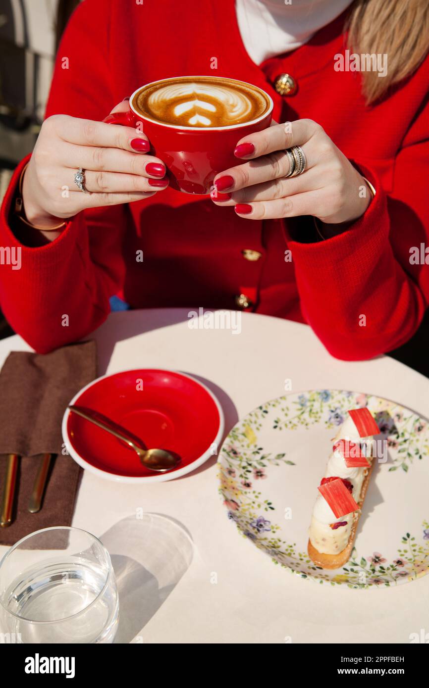 Stylish woman with red nails drinks cappuccino coffee and eats cake in