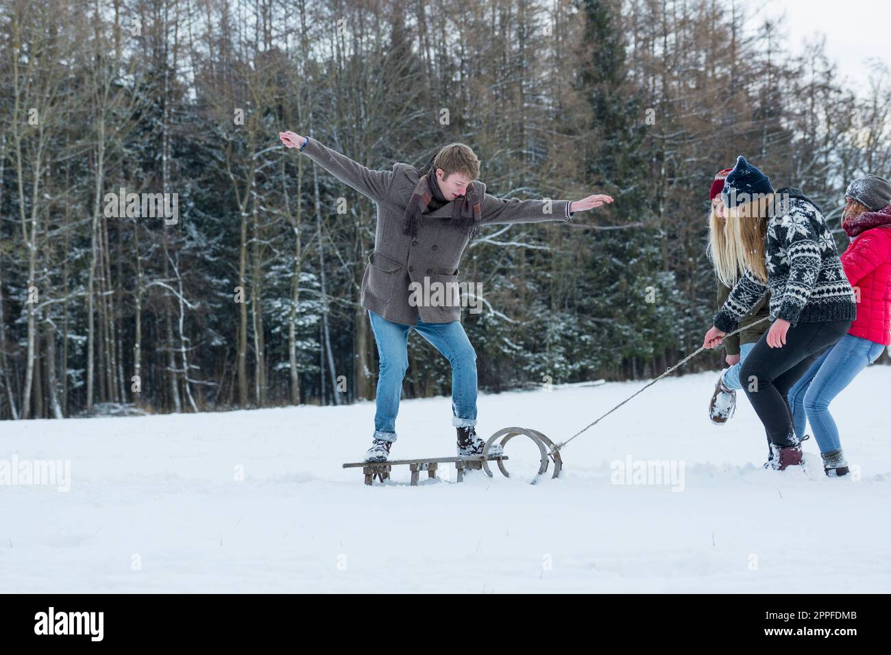 Three girls pulling young man standing on sled in snowy landscape