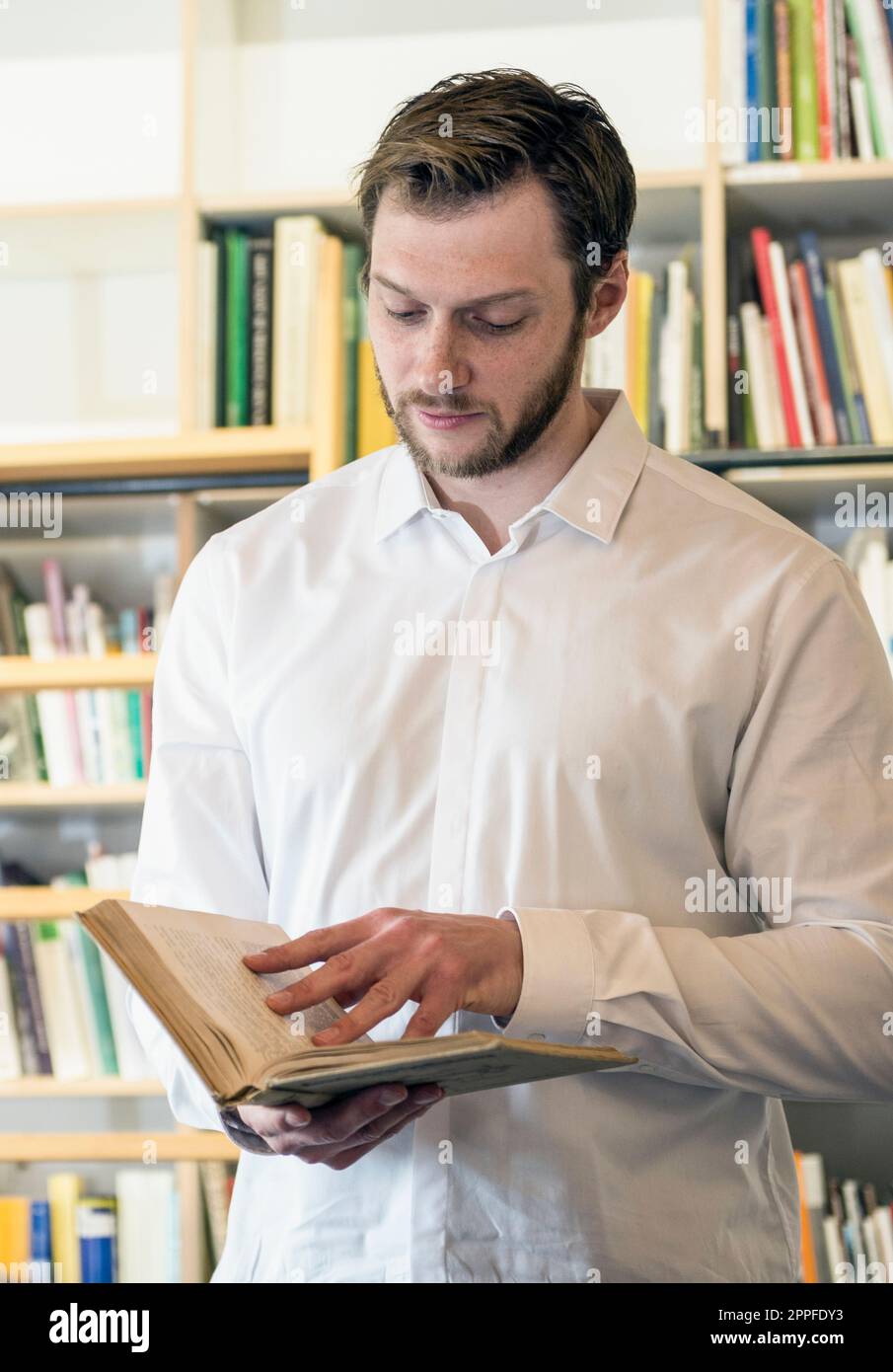 Mid adult businessman reading book in an office in front of bookshelf, Bavaria, Germany Stock Photo