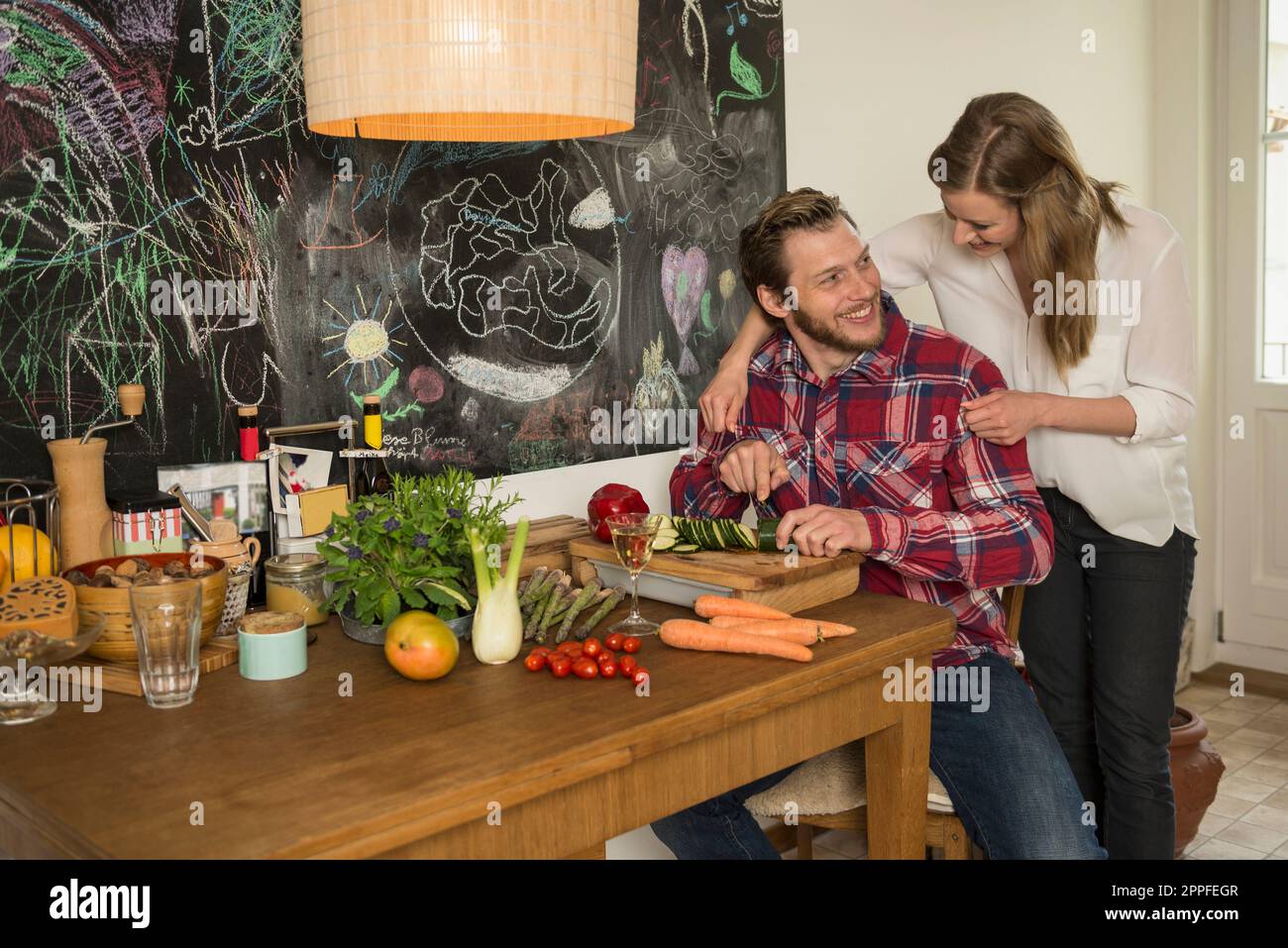 Man slicing vegetables on chopping board and his wife hugging from behind, Munich, Bavaria, Germany Stock Photo