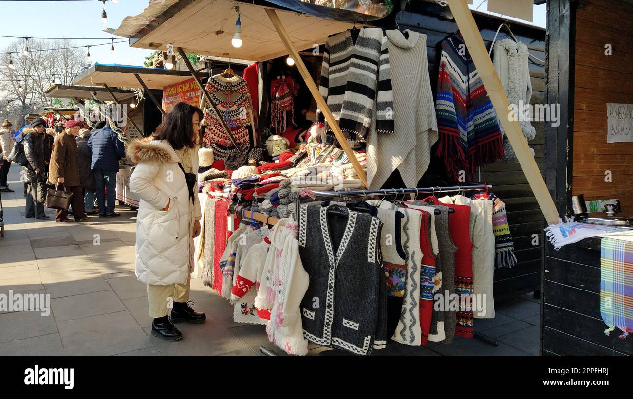 A woman of Asian appearance looks at goods in a street shop. On the window of the stall are beautiful knitted sweaters, vests, coats. Souvenir fair Stock Photo
