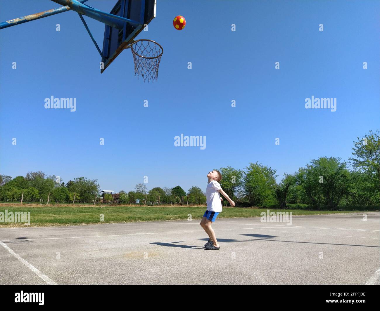The boy throws the ball into the basketball net. The child plays with a ball on the playground. Free space for text. Blue sky in the background. Child with blond hair. Stock Photo