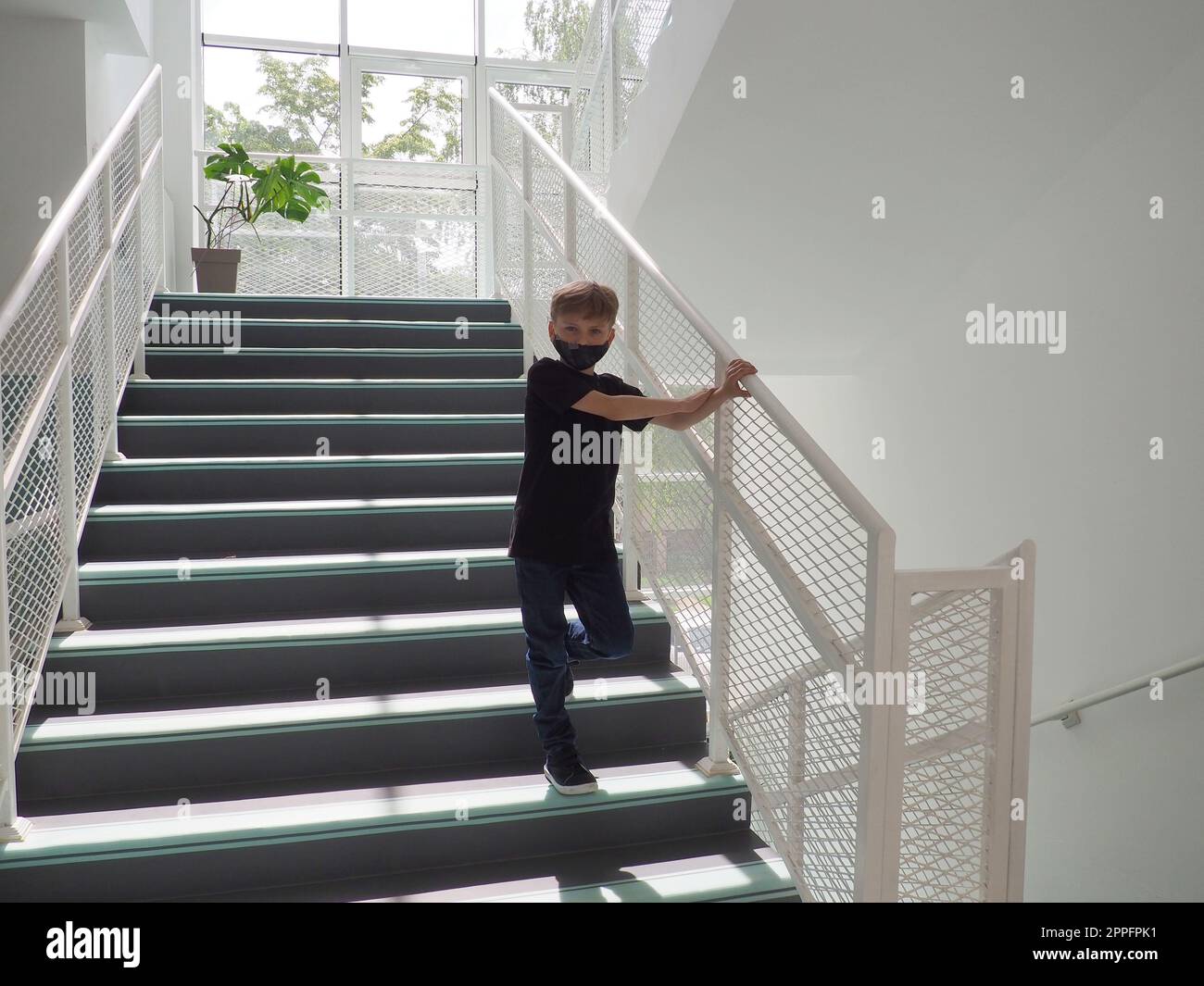 A blond boy 8 years old in black clothes and a mask stands on a white school ladder. The child looks at the camera and holds the handrail with his hand. School concept. Internal interior. Stock Photo