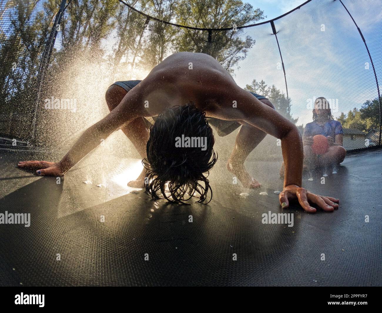 Backlit shot of boy putting head in sprinkler on trampoline in summer Stock Photo