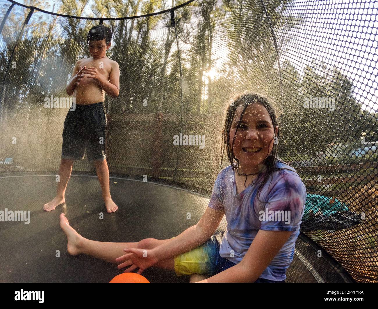 Smiling child on trampoline with brother in background and water spray Stock Photo