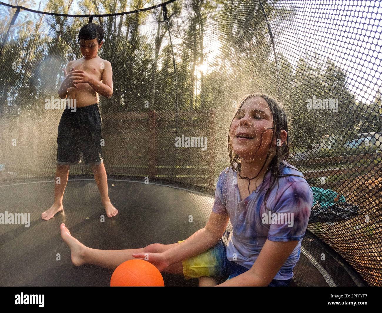 Child happily cooling off with sprinkler on trampoline at sunset Stock Photo