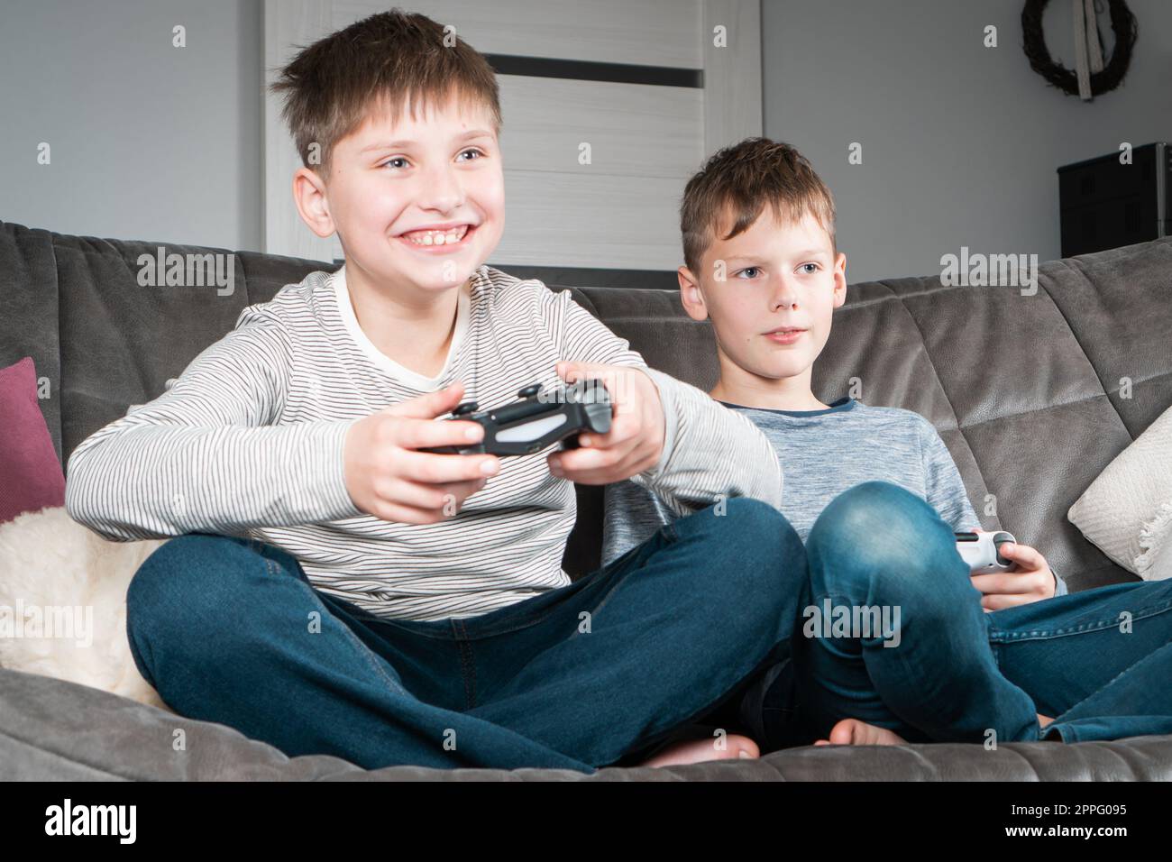 Portrait of two glad teenage boys sitting on grey sofa at home, holding gaming controller joystick, playing videogames. Stock Photo