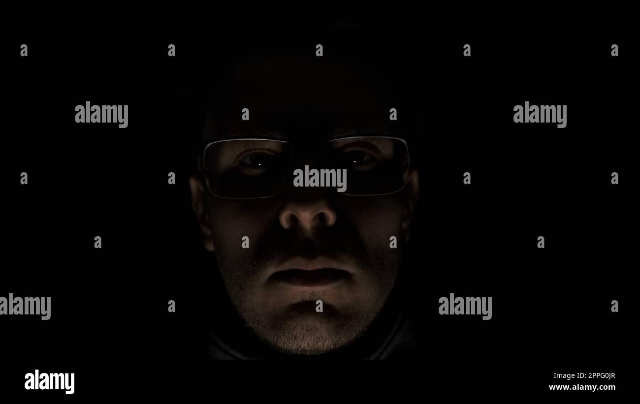 the man in glasses looking in camera on a black background in low key anger, psychopath emotions. Horror maniac face in pulsing light. Mental disorder of mind. Stock Photo
