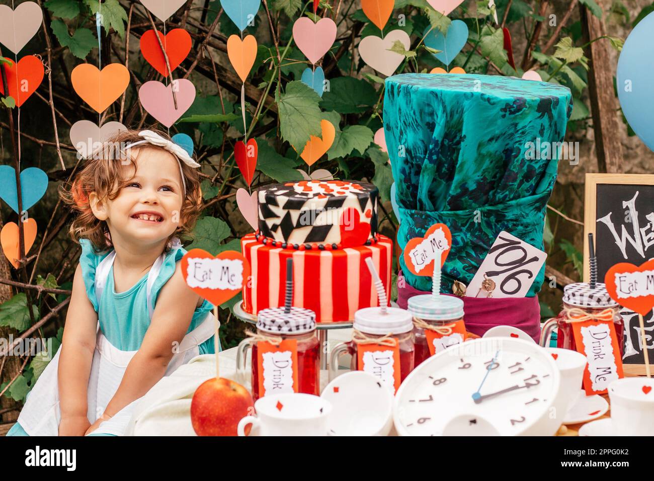 Little smiling girl dressed as Alice in Wonderland near table with fairy tale props. Cake, Hatter hat, clock, card suits Stock Photo