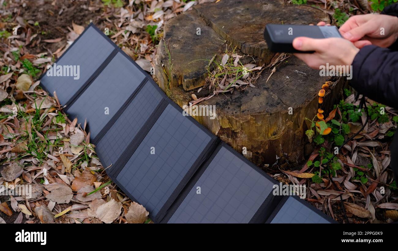 Hands plug in portable solar panel charging power bank by mutlipurpose cable, smartphone, watches, laptops nature background. Clean energy for using in camping or at home when NO electricity. Stock Photo