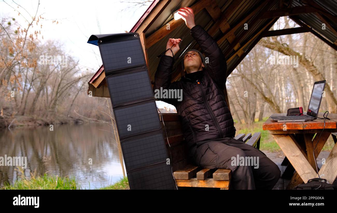 Man work on laptop charging from portable solar panel utdoor in alclove near the river. Clean energy for using in camping. Work with gadgets outdoor with portable solar panels. Stock Photo