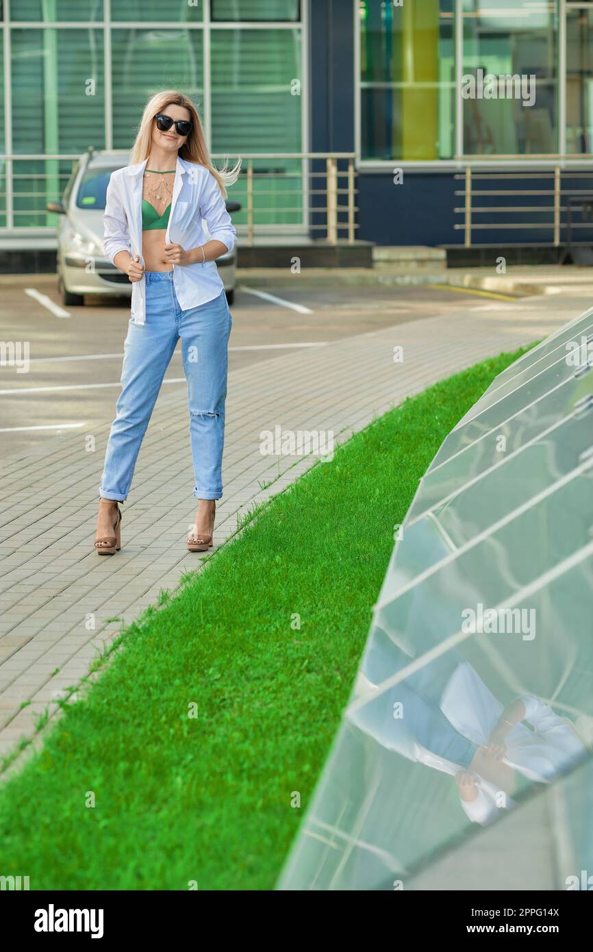 Pretty lady in blue jeans and white leather jacket near modern office building Stock Photo