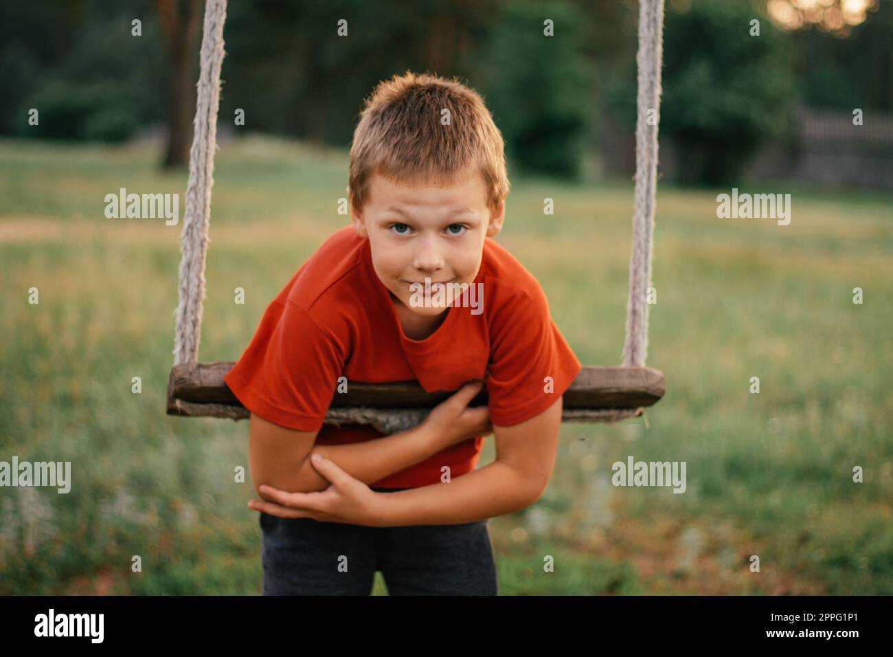 Barefoot boy 7-10 in red t-shirt stands leaning on rope swing in countryside and looks into frame smiling in rays of sun Stock Photo