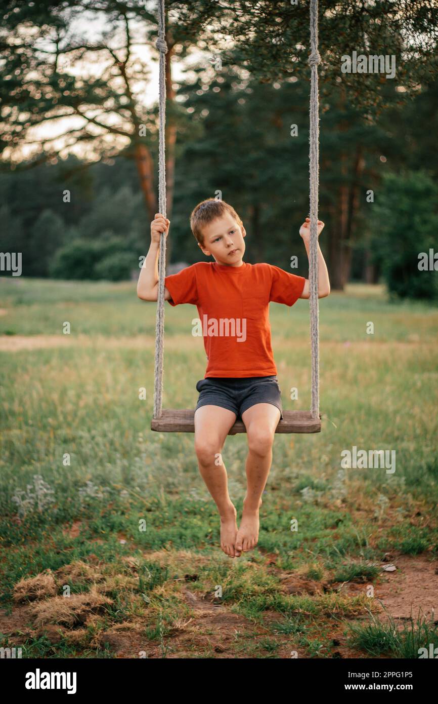 Barefoot boy 7-10 in red t-shirt with bruised legs sitting on rope swing in countryside among pines. Vertical Stock Photo