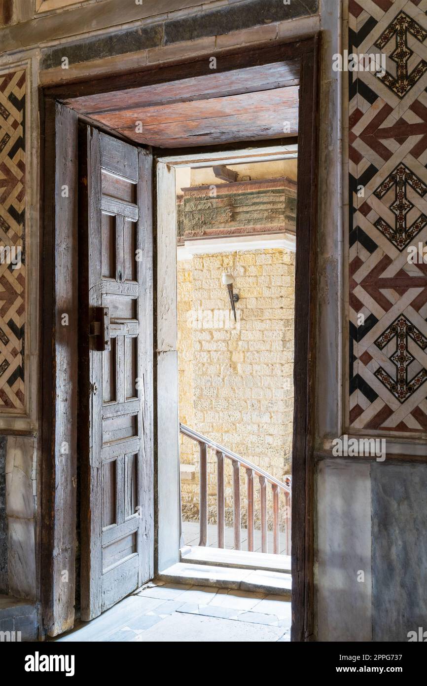 Wooden aged ornate opened door leading to a passage with bright light, color decorated marble wall, and marble floor Stock Photo