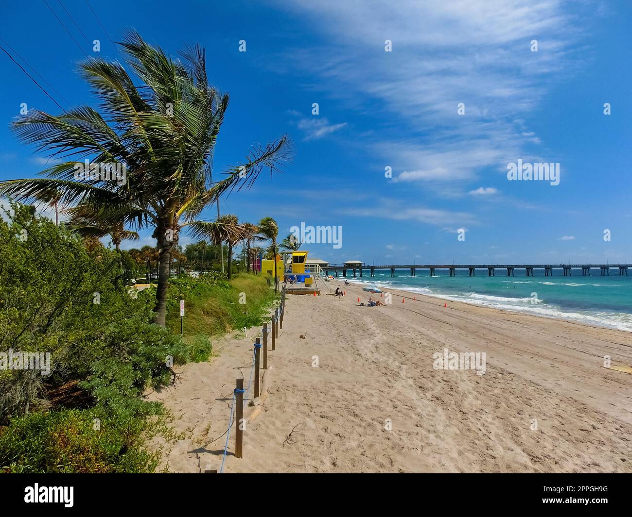 Long distance view of the sunny and tropical blue water shoreline with people, foliage, boats and buildings in the background seen from above the water. Stock Photo