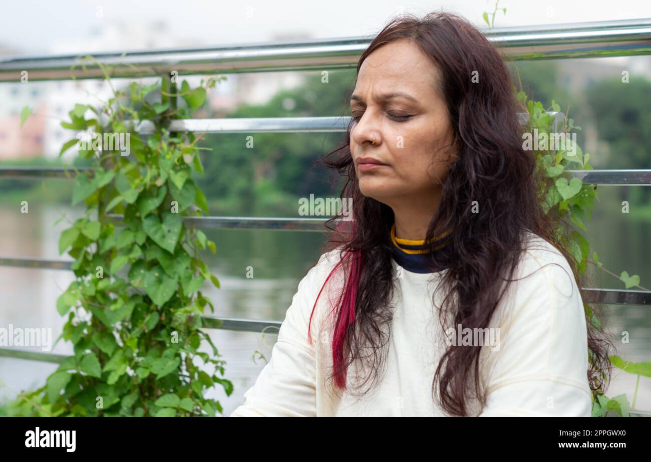 Active Mid Adult Woman in meditation sitting with eyes closed concentrate in a public park against garden leaves in the background. Front view. Head and Shoulder Shot. Close up Portrait. Stock Photo