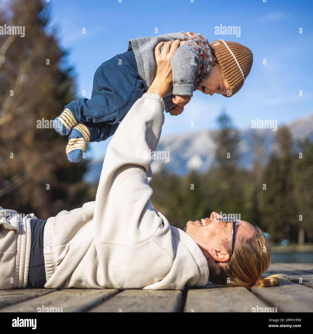 Happy family. Young mother playing with her baby boy infant oudoors on sunny autumn day. Portrait of mom and little son on wooden platform by lake. Positive human emotions, feelings, joy. Stock Photo