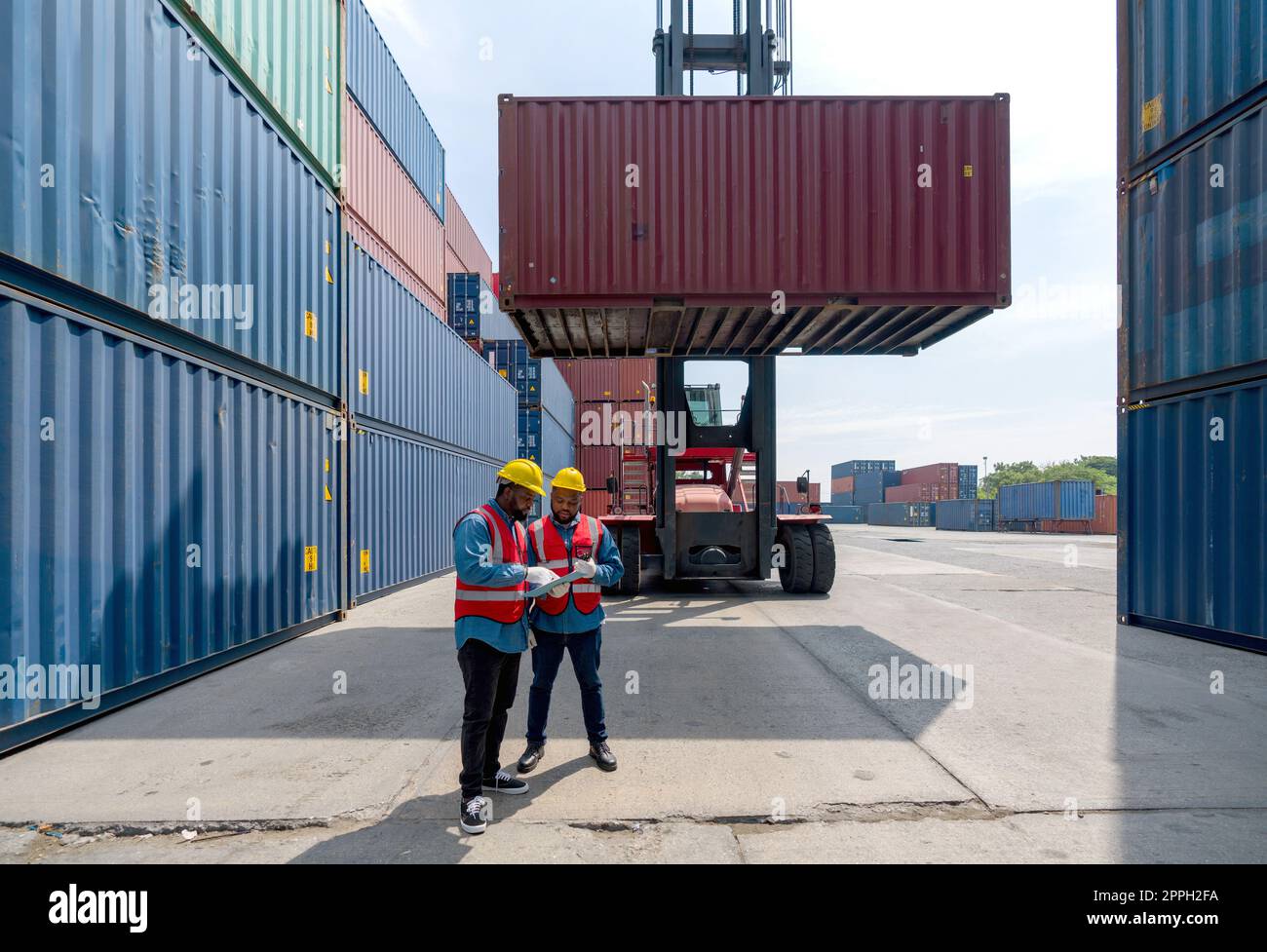 Two short black hair man with moustache and beard dressed in hardhat, safety vest and protective glove working during the day under sunlight. There are heavy duty container forklift in the work area. Stock Photo