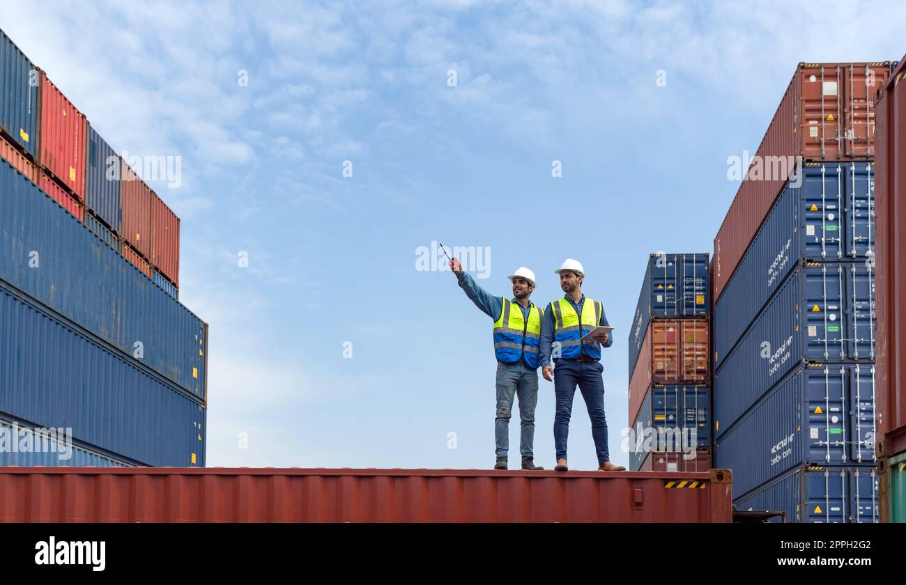 Shipment worker use a walkietalkie to point to container storage location, explain to colleague about planning for next shipment. Young worker with safety vest holding tablet computer stand beside him Stock Photo