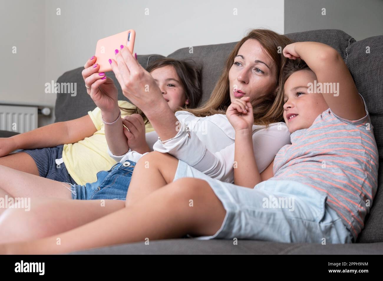 Happy mother with her kids are making a selfie or video call to father or relatives in a sofa. Concept of technology, new generation, family, connection, parenthood. Stock Photo