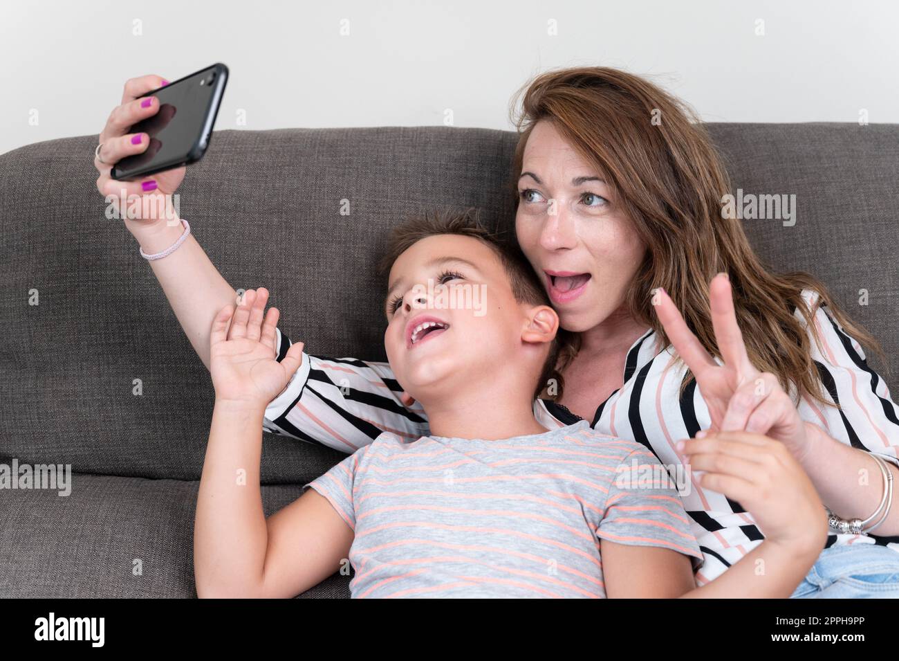 Cheerful mother holding smart phone making selfie with her little son showing victory sign. mom and kid boy sit on couch using gadget having fun taking self-portrait spend time together at home . Stock Photo