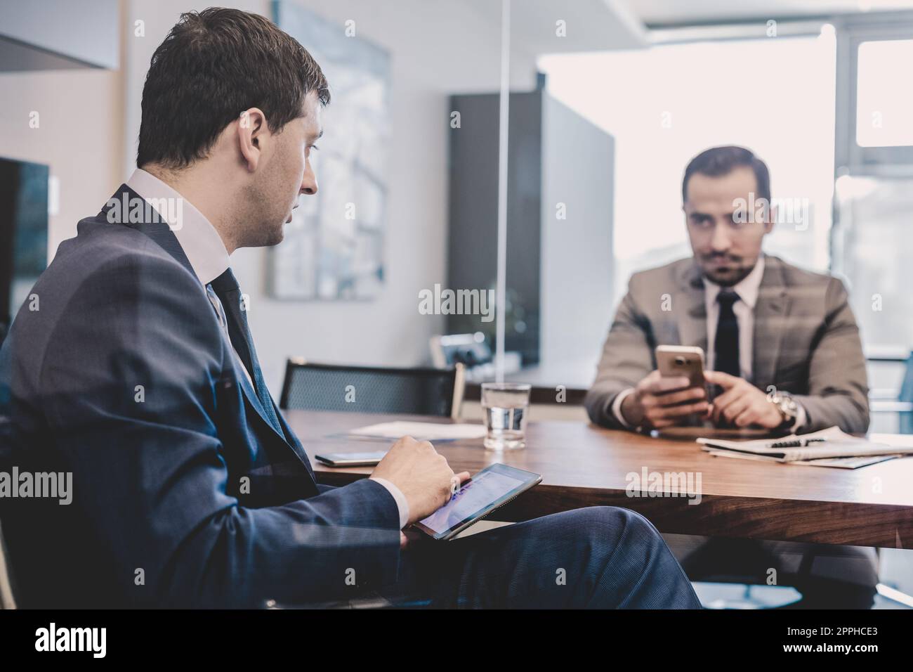 Two businessmen using smart phones and touchpad at meeting. Stock Photo